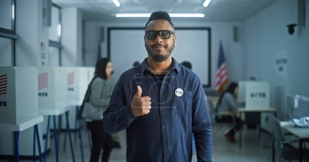 Téléchargez les photos : Portrait de l'homme afro-américain, électeur des États-Unis d'Amérique. L'homme avec badge se tient dans un bureau de vote moderne, pose, regarde la caméra. Contexte avec isoloirs. Jour du scrutin aux États-Unis. - en image libre de droit