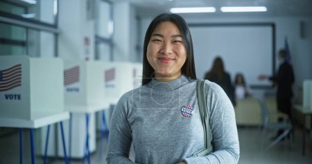 Téléchargez les photos : Portrait de femme asiatique, électrice des États-Unis d'Amérique. Femme avec badge se tient dans un bureau de vote moderne, pose, sourit, regarde la caméra. Contexte avec isoloirs. Concept du devoir civique. - en image libre de droit