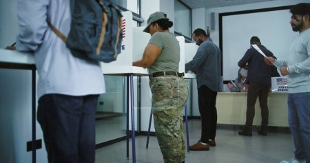 American female soldier stands at voting booth in polling station office. National Election Day in United States. Political races of US presidential candidates. Concept of civic duty and patriotism.