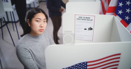 Photo for Dolly shot of Asian female voter choosing presidential candidate to vote for in voting booth at polling station, then looking at camera. US citizen during National Election Day in the United States. - Royalty Free Image