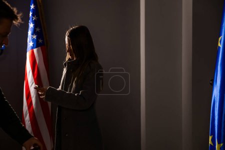 Silhouette of a female politician with a smartphone in her hands stands between the flags of the USA and the EU