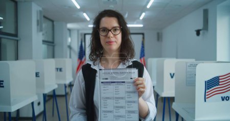 Foto de Mujer votante o oficial de votación estadounidense habla en cámara, muestra papeleta de papel, pide votación. Día Nacional de Elecciones en los Estados Unidos. Cabinas de votación en el centro de votación. Deber cívico y patriotismo - Imagen libre de derechos