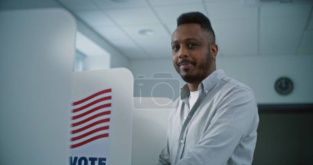 Photo for African American man fills out paper ballot in voting booth, than smiles and looks at camera. US citizen or male voter at polling station during National Election Day in the United States of America. - Royalty Free Image