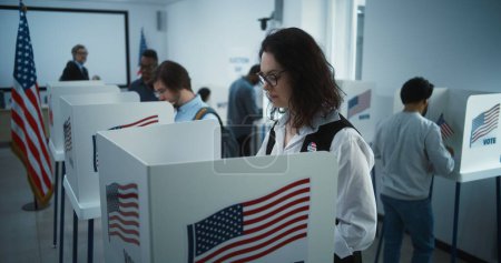 Téléchargez les photos : Une femme à lunettes vote dans un bureau de vote. Journée nationale des élections aux États-Unis. Courses politiques des candidats à la présidence américaine. Devoir civique et patriotisme. Au ralenti. Dolly shot. - en image libre de droit