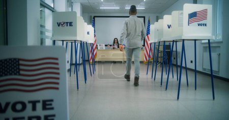 Foto de Ciudadano estadounidense, votante afroamericano camina hacia oficial electoral femenino para registrarse en el centro de votación, toma el boletín de papel para votar. Día Nacional de Elecciones en los Estados Unidos. - Imagen libre de derechos