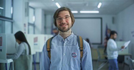 Téléchargez les photos : Portrait d'un homme caucasien, électeur des États-Unis d'Amérique. L'homme avec badge se tient dans un bureau de vote moderne, pose, sourit, regarde la caméra. Contexte avec isoloirs. Concept du devoir civique. - en image libre de droit