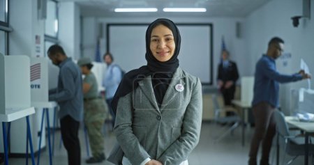 Foto de La mujer se encuentra en un moderno centro de votación, posa, sonríe y mira a la cámara. Retrato de la mujer árabe, votante de las elecciones de los Estados Unidos de América. Antecedentes con mesas de votación. Concepto de deber cívico. - Imagen libre de derechos