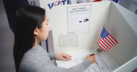 Téléchargez les photos : Une femme asiatique vient à l'isoloir au bureau de vote, fait un choix et remplit un bulletin papier. Citoyen américain lors du jour national des élections aux États-Unis d'Amérique. Devoir civique et patriotisme. - en image libre de droit