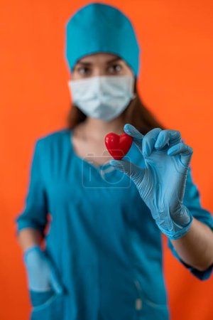 Téléchargez les photos : Médecin ou infirmière en uniforme bleu avec des gants de masque tenant un petit cœur rouge dans la salle d'opération hôpital. Concept de médecine. Opérationnel. Un symbole d'amour et de vie - en image libre de droit