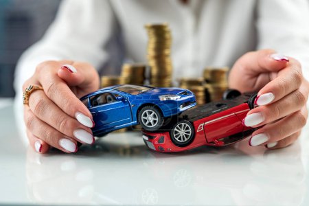Foto de Manos de una mujer sosteniendo dos modelos de coche simulando un accidente cerca de una montaña de monedas. Seguro de auto. Accidente de coche conducción seguridad - Imagen libre de derechos