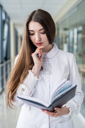 Photo for Smiling female doctor holding a book or magazine and walking down the med corridor to examine patients. Portrait of a female doctor. Medicine saves lives. A beautiful nurse - Royalty Free Image