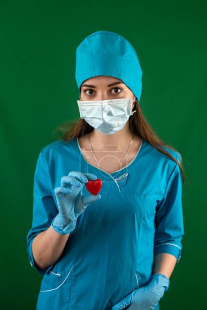 Téléchargez les photos : Médecin ou infirmière en uniforme bleu avec des gants de masque tenant un petit cœur rouge dans la salle d'opération hôpital. Concept de médecine. Opérationnel. Un symbole d'amour et de vie - en image libre de droit