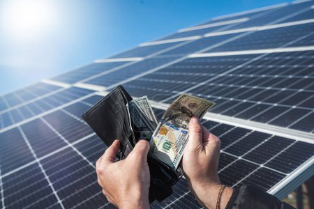 Foto de Money dollars in a wallet holding hands over a solar panel. Banknotes on the panel. Concept of cheap solar energy. - Imagen libre de derechos