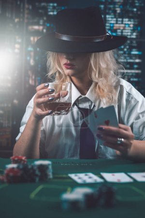 Photo for Attractive girl in shirt and hat drinking whiskey and looking at poker cards in casino. girl player makes a bet. poker in the casino - Royalty Free Image