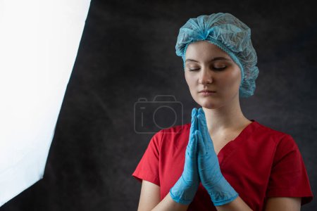 Photo for Young feamle doctor in uniform prays for God's, isolated on dark, studio shot. Concept about hopes that everything will be better. - Royalty Free Image