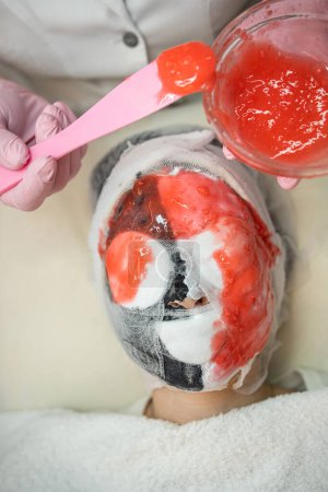 Téléchargez les photos : Cosmétologue appliquant un masque d'alginate rouge à visage pour sa cliente dans un salon de beauté. Concept de traitement anti-âge - en image libre de droit