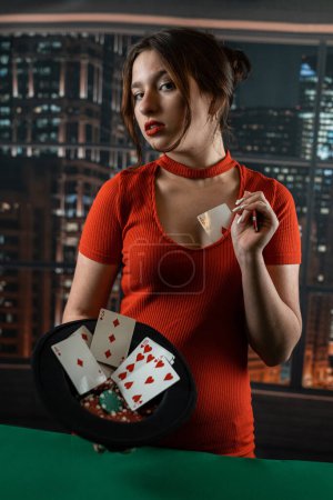 Photo for Beautiful young girl in a red dress holding a hat in which card and colored chips near the poker table. poker concept. woman - Royalty Free Image