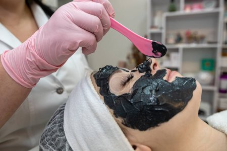 Téléchargez les photos : Cosmétologue appliquer masque crème noire sur le visage de son client. Soins spa, soins du corps - en image libre de droit