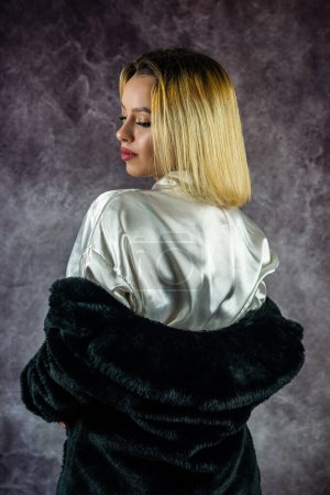 Téléchargez les photos : Jeune femme dans une belle robe de soie légère avec un manteau de fourrure noir chaud sur le dessus tourne la tête. isolé sur fond fumé. concept portrait féminin - en image libre de droit