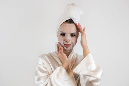 Téléchargez les photos : Jeune femme en serviette blanche applique un masque hydratant en tissu blanc isolé sur fond blanc, procédure anti-âge - en image libre de droit