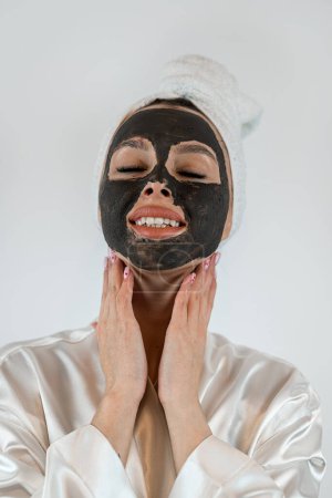 Téléchargez les photos : Belle jeune femme avec masque facial en argile noire pour une peau fraîche et propre, isolée sur du blanc. Apprécie les soins de beauté - en image libre de droit