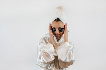 Hübsche junge Frau in Handtuch mit schwarzem Kollagenpflaster unter ihrem Auge, isoliert auf weiß. Hautpflegekonzept.