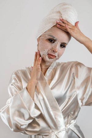 Téléchargez les photos : Jeune femme en serviette blanche applique un masque hydratant en tissu blanc isolé sur fond blanc, procédure anti-âge - en image libre de droit