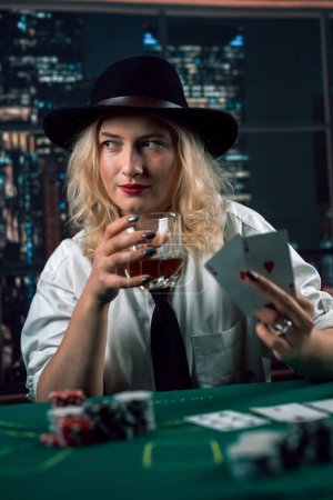 attractive girl in shirt and hat drinking whiskey and looking at poker cards in casino. girl player makes a bet. poker in the casino