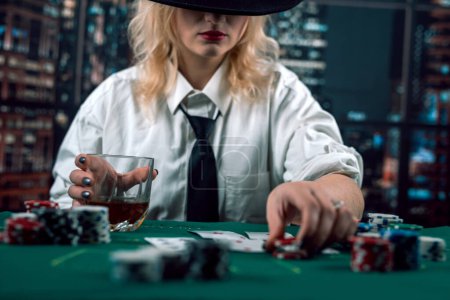 Photo for Attractive girl in shirt and hat drinking whiskey and looking at poker cards in casino. girl player makes a bet. poker in the casino - Royalty Free Image