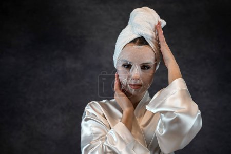 Téléchargez les photos : Femme porter serviette blanche appliquer feuille de tissu masque cosmétique pour nettoyer la peau du visage soins isolés sur noir. Concept de soins de la peau. procédure pour la peau pour rajeunir et éliminer les défauts - en image libre de droit