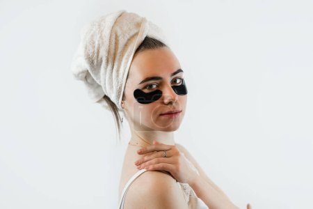 caucasian girl in towel with black patches under eyes isoalted on white.   Anti wrinkle collagen eye patches. Healthy skin 