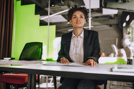 Téléchargez les photos : Portrait taille haute d'une belle employée afro-américaine calme assise seule à la table - en image libre de droit