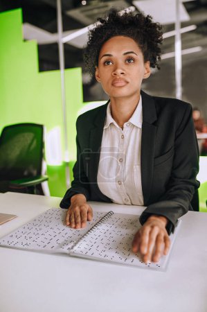 Téléchargez les photos : Jeune employé de bureau assis à la table touchant des points soulevés dans le carnet ouvert relié en spirale - en image libre de droit