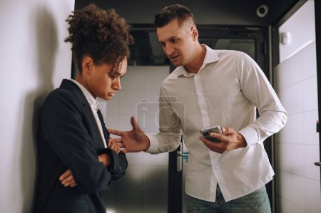 Photo for Clarification. Caucasian middle aged man with smartphone talking looking at sad mulatta woman standing sideways to camera in office - Royalty Free Image
