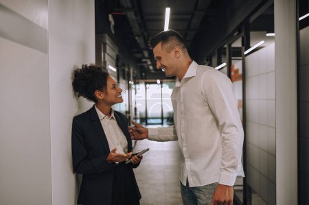 Positive communication. Young adult mulatto woman with smartphone and caucasian man touching shoulder standing sideways to camera smiling communicating