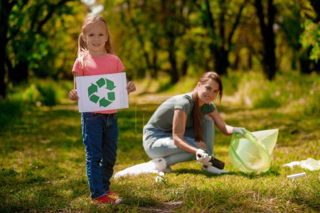 Photo for Eco activity. Blonde girl with eco symbol and her mom gathering garbage in the park - Royalty Free Image