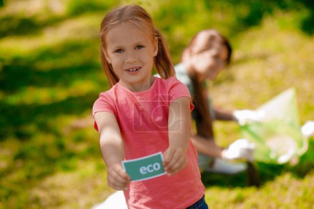 Photo for Eco activity. Blonde girl with eco symbol and her mom gathering garbage in the park - Royalty Free Image