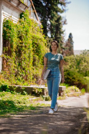 Photo for Freelancer. Young woman with a laptop in hands walking in the park - Royalty Free Image