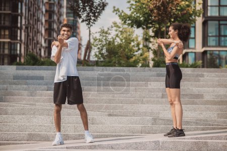 Photo for Smiling young sporty guy and a cheerful fit girl doing a shoulder stretch exercise outdoors - Royalty Free Image