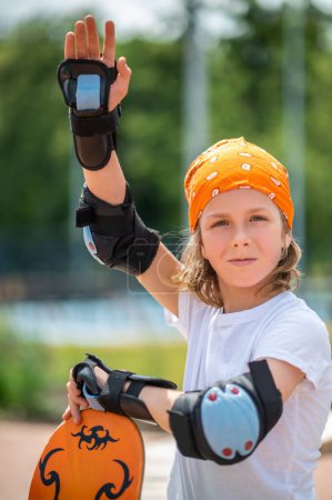 Photo for Kid with the wrist guards and elbow pads on the arms holding his skateboard and waving at someone - Royalty Free Image