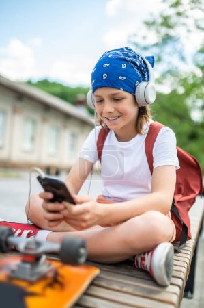 Foto de Smiling preadolescent boy with the backpack over the shoulders sitting on the bench while looking at the smartphone touch screen - Imagen libre de derechos