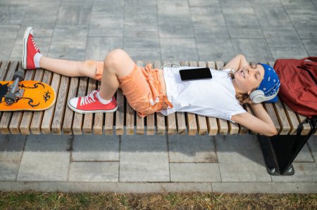 Foto de Joyful preadolescent boy lying down on his back with closed eyes and listening to music through his wireless headphones - Imagen libre de derechos