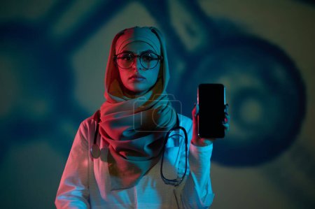 Foto de Médico en línea. Doctora joven seria en hiyab con un teléfono en la mano - Imagen libre de derechos