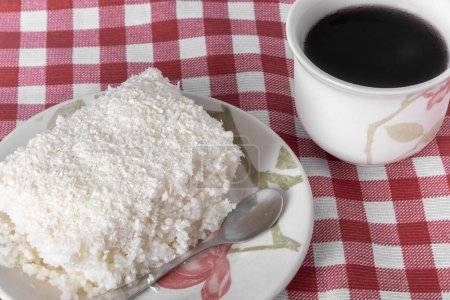 Photo for Piece of couscous in porcelain saucer on checkered tablecloth, typical Brazilian dish, made with tapioca, milk, sugar and coconut milk. With cup of coffee. - Royalty Free Image