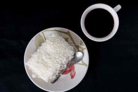 Photo for Piece of couscous in porcelain saucer on black background, typical Brazilian dish, made with tapioca, milk, sugar and coconut milk. With cup of coffee. Top view. - Royalty Free Image