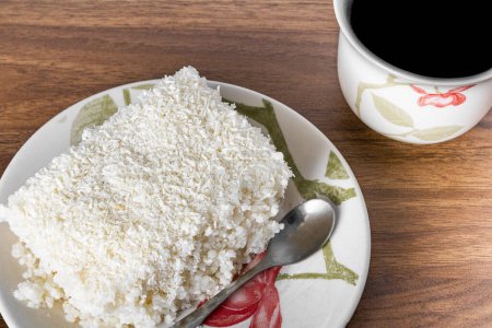 Photo for Piece of couscous in porcelain saucer on wooden table, typical Brazilian dish, made with tapioca, milk, sugar and coconut milk. With coffee cup. - Royalty Free Image