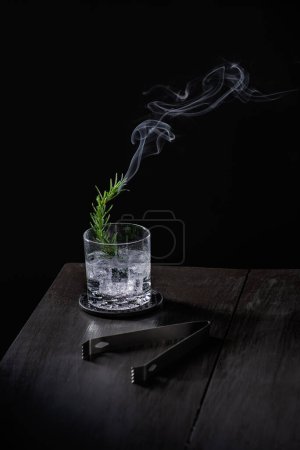 Téléchargez les photos : Branche de romarin éclairée de fumée à l'intérieur d'une boisson mezcal avec tequila sur glace, pinces à glace sur une table en bois, personne - en image libre de droit