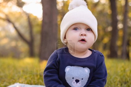 Photo for Baby in knitted hat sits on grass in Park against background of autumn trees. children's clothing. walk outdoor. High quality photo - Royalty Free Image