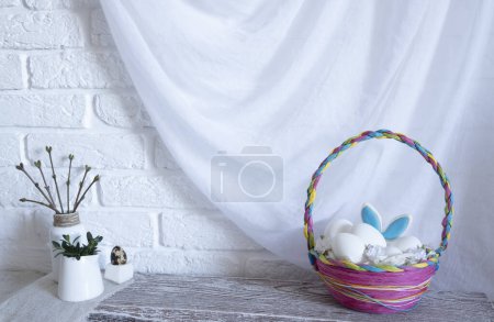 Téléchargez les photos : Un panier de Pâques en osier coloré avec des œufs est sur la table dans la cuisine. Les oreilles d'un lièvre sortent du panier. Photo de haute qualité - en image libre de droit
