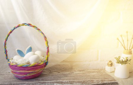 Téléchargez les photos : Un panier de Pâques en osier coloré avec des œufs est sur la table dans la cuisine. Les oreilles d'un lièvre sortent du panier. Photo de haute qualité - en image libre de droit